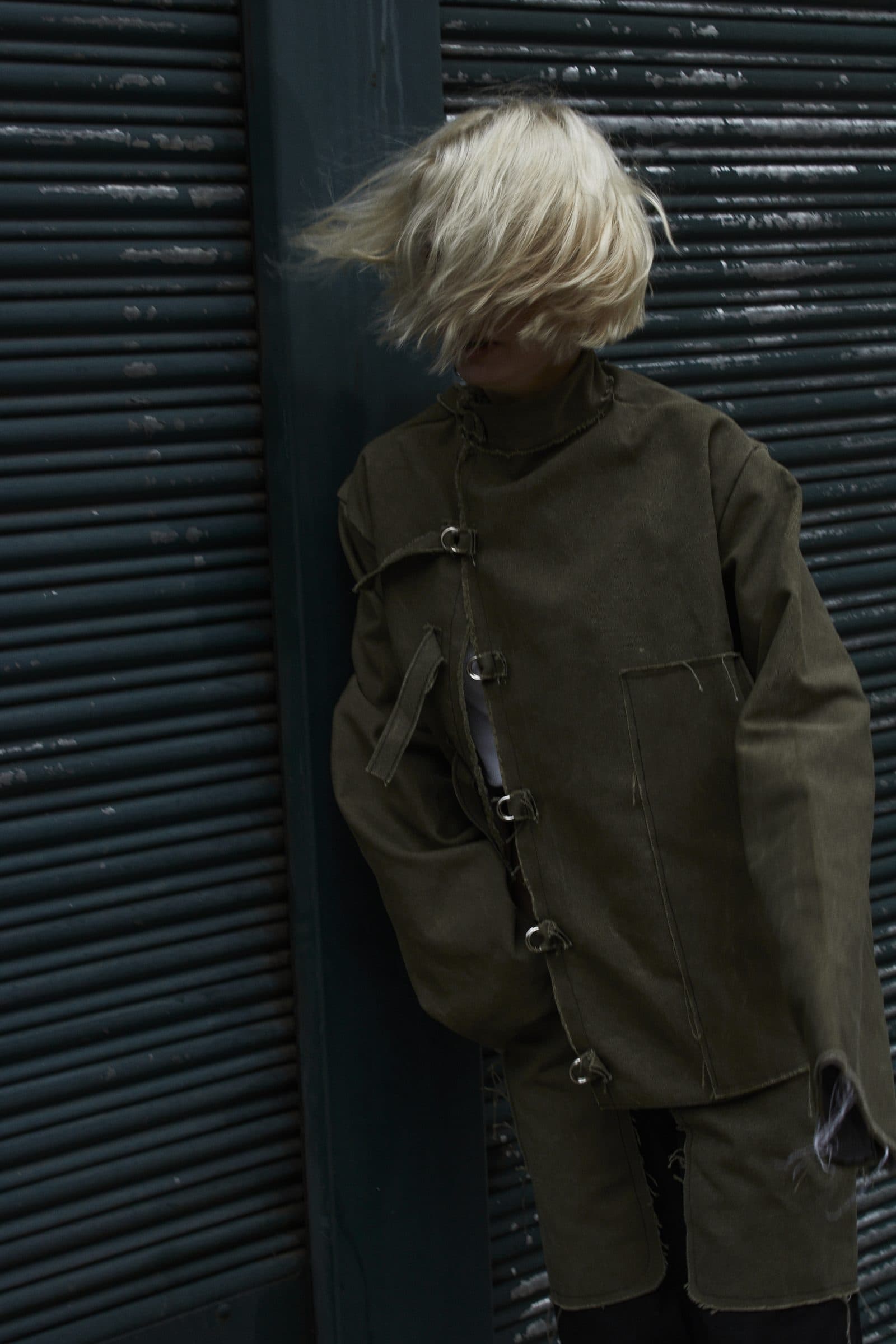 AKIN jacket in green tapestry, worn in front of a blue rolling shutter, hair in motion