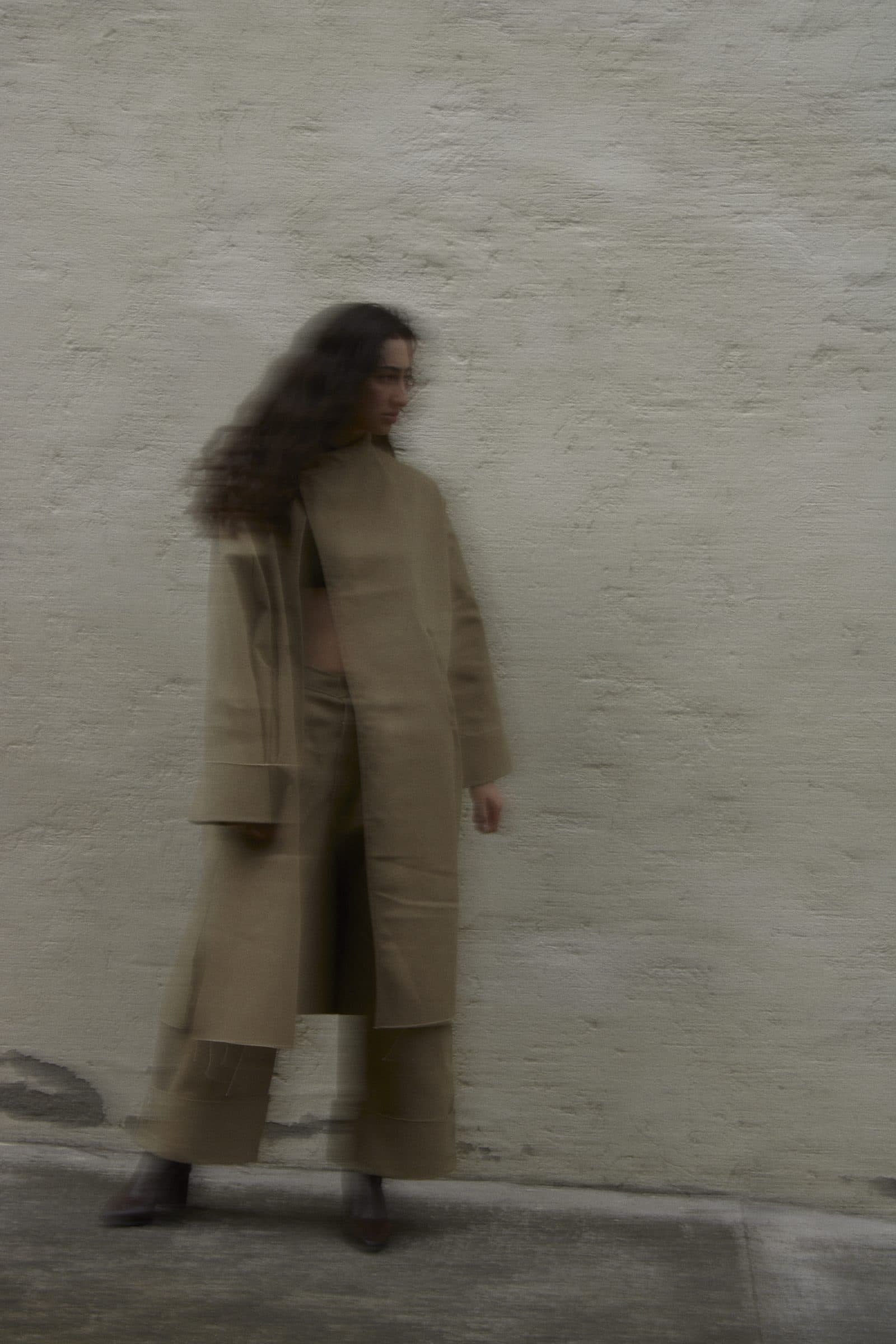 A model in the FRAME coat and wide trousers, turning in motion against a pale wall